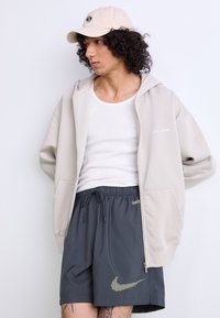 Beige zippered hættejakke med logo, hvid tanktop, mørkegrå shorts med et gult Nike-logo og en beige cap med logo.