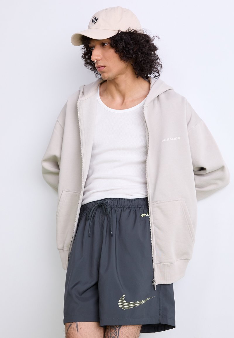 Felpa beige con zip e logo, canotta bianca, pantaloni corti grigi scuri con logo giallo Nike, e cappellino beige con logo.