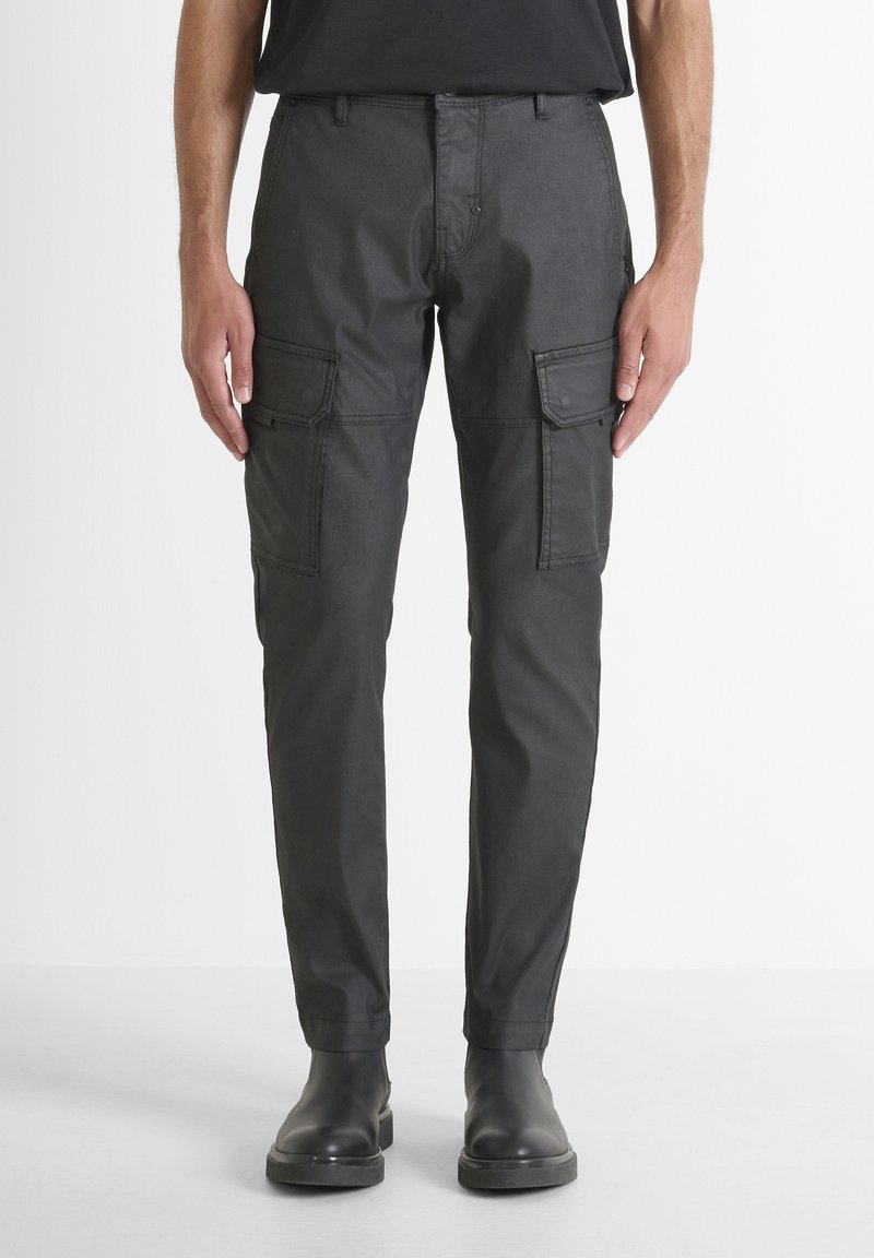 Uomo che indossa pantaloni cargo grigi scuri con tasche laterali e stivali neri, in piedi di fronte a uno sfondo semplice chiaro.