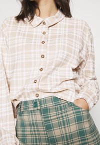 Béžová károvaná košile na knoflíky s límečkem a dřevěnými knoflíky, doplněná zelenými károvanými kalhotami s vysokým pasem a kontrastním detailem na knoflíku.
