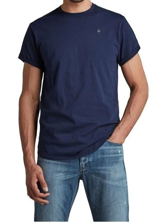 Mann trägt ein schlichtes dunkelblaues Kurzarm-T-Shirt mit hochgekrempelten Ärmeln und hellblaue Jeans, eine Hand in der Tasche, vor weißem Hintergrund.