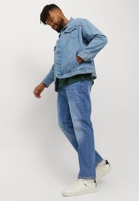 Lichtblauwe denimjack met twee zakken, gedragen over een donkergroen shirt, gecombineerd met lichtblauwe jeans en witte sneakers. Zachte textuur.