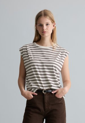 Jeune femme aux cheveux blonds porte un haut sans manches blanc rayé de marron et un pantalon marron foncé, debout les mains dans les poches.