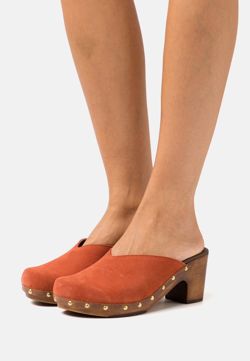 Femme portant des sabots en daim couleur rouille avec des talons blocks en bois et des clous dorés le long du bord de la semelle.