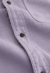 Lavendlivärvi nööpidega särk, millel on tekstuuriga kangas, nähtavad õmblused ja pruunid nööbid eesmisel plikal.