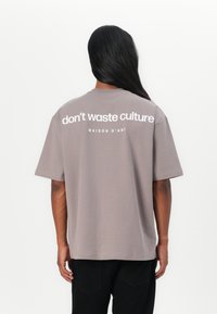 Camiseta de algodón gris con mangas cortas y cuello redondo. La parte trasera presenta texto blanco: "no desperdicies la cultura MAISON D'ART." Diseño minimalista.
