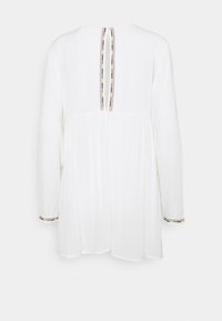 Robe blanche à manches longues avec une ouverture verticale en forme de trou de serrure dans le dos, ornée d'un bord cousu noir et or aux bords et poignets.
