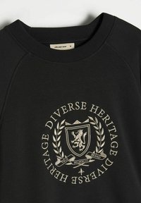 Czarna bluza z ribbnowym dekoltem, z kremowym haftowanym herbem i napisem: "Różnorodne Dziedzictwo," otoczonym gałązkami wawrzynu.