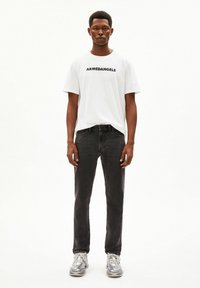 Jonge man staand, gekleed in een witte T-shirt met "ARMEDANGELS" opschrift, zwarte spijkerbroek en grijze sneakers, tegen een effen witte achtergrond.