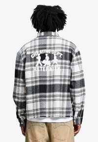 Chemise en flanelle noir et blanc avec un motif à carreaux, arborant une impression graphique disant "CREATIVE DEPT. RETENITY" dans le dos.