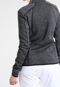 Woman wearing a fitted dark gray textured jacket with black trim and white pants, shown from behind against a plain background.