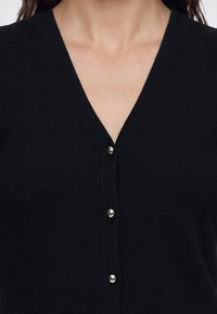 Femme portant un cardigan noir à col en V avec quatre boutons argentés visibles.