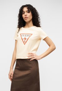 Κρεμ κοντομάνικο t-shirt με πορτοκαλί λογότυπο σε σχήμα τριγώνου. Συνδυάζεται με καφέ φούστα, με λεία υφή και εφαρμοστή γραμμή.