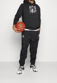 Schwarze Kapuzenjacke mit weißem Nets-Logo, kombiniert mit schwarzen Trainingshosen mit einem weißen Streifen und Logo sowie schwarz-weißen Sportschuhen.