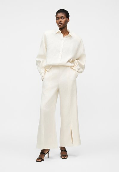 Γυναίκα που φοράει μια υπερμεγέθη κρεμ πουκάμισο με κουμπιά και παντελόνια με φαρδιά μπατζάκια, συνδυασμένα με μαύρα σανδάλια με ψηλό τακούνι, στέκεται μπροστά σε λευκό φόντο.