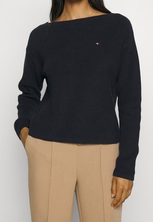 Femme portant un pull noir côtelé avec un petit logo rouge, blanc et bleu, associé à un pantalon beige, debout avec les mains dans les poches.