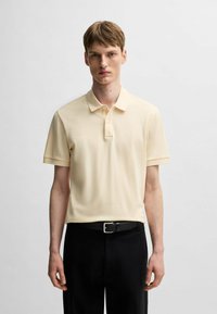 Polo in morbido tessuto giallo chiaro, maniche corte, patta con tre bottoni e ricamo del logo discreto. Indossato con cintura nera e pantaloni.
