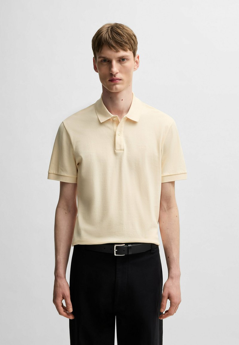 Polo in morbido tessuto giallo chiaro, maniche corte, patta con tre bottoni e ricamo del logo discreto. Indossato con cintura nera e pantaloni.
