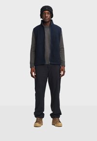 Gilet in pile blu navy con maglione a maniche lunghe grigio in maglia, pantaloni cargo neri e stivali color sabbia. Un berretto di lana completa l'outfit.