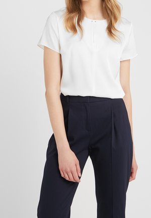 Vrouw draagt een witte blouse met korte mouwen, ingestopt in een donkerblauwe, op maat gemaakte, hooggesneden broek, tegen een effen achtergrond.