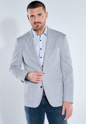 Mann trägt hellgrauen strukturierten Blazer über hellblauem Hemd mit Knöpfen und dunklen Jeans, richtet den vorderen Knopf des Blazers, neutraler Hintergrund.