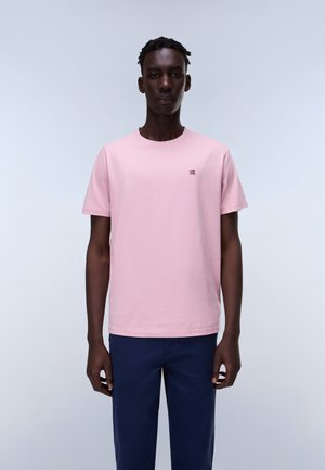 Uomo in piedi rivolto in avanti che indossa una T-shirt rosa chiaro a maniche corte con un piccolo logo a forma di bandiera e pantaloni blu scuro.