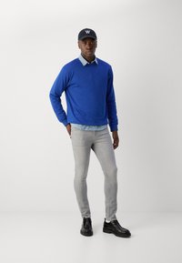 Sudadera azul superpuesta sobre una camisa de cuadros azul claro, combinada con jeans slim-fit gris claro y zapatos negros. Gorra negra con logo blanco.