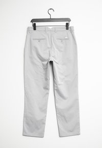 Pantalons gris clair en tissu avec une texture lisse. Équipés de deux poches arrière avec fermetures à bouton et de passants de ceinture. Suspendus à un cintre noir à pince.