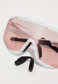 Lunettes de sport teintées de rose avec une monture blanche, présentant un design angulaire, des plaquettes en caoutchouc pour le nez et des embouts de branches noirs ajustables.