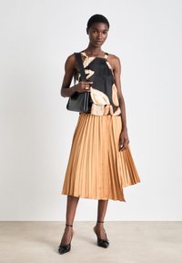 Blouse sans manches noire avec un motif floral, jupe plissée beige et sac à main noir. Le mannequin porte des ballerines noires à bouts pointus avec des brides à la cheville.