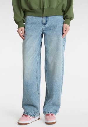 Osoba nosząca luźne, jasnoniebieskie dżinsy, zieloną bluzę z kapturem na zamek i różowe trampki na jednolitym białym tle.