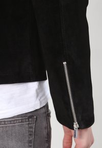 Giacca in suede nero dal design elegante, con una zip laterale argentata e una finitura leggermente testurizzata. La giacca ha maniche su misura.
