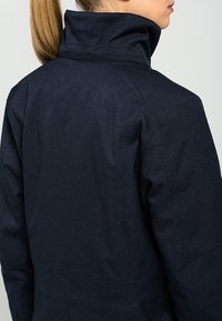 Dark blue jacket with a high collar, made from textured material. Features stitching details and a subtle logo on the collar.