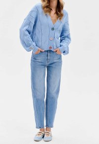 Ljusblå stickad kofta med v-ringning och strukturerade mönster, med tre knappar i olika färger, kombinerad med högmidjade jeans.