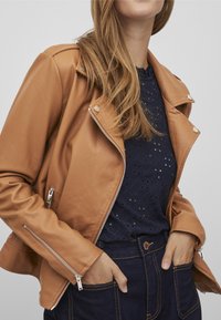 Beżowa kurtka motocyklowa z ekoskóry z srebrnymi detalami, zamykanymi kieszeniami i lekko teksturowanym wykończeniem, na ciemnoniebieskiej bluzce z detalami w kształcie oczek.