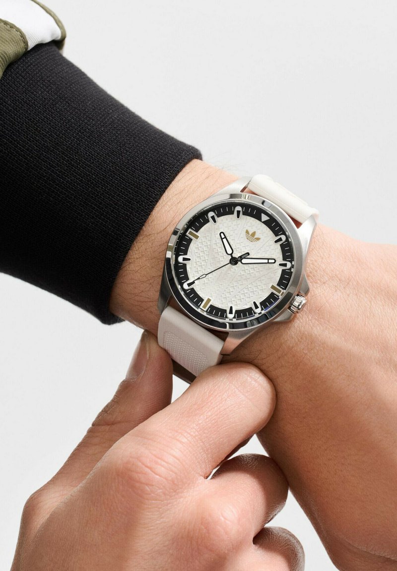 Sportinis laikrodis su baltu silikoniniu dirželiu, nerūdijančio plieno korpusu, juodu rėmeliu ir tekstūruotu baltu ciferblatu su ryškiais valandų žymekliais ir rodyklėmis.