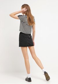 Černá džínová mini sukně v kombinaci s černobílým pruhovaným topem s krátkými rukávy. Modelka je otočená zády, má na sobě černé boty a bílé ponožky.