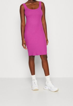 Kvinde iført en ærmeløs, knælængde, ribbet lilla kjole kombineret med hvide sokker og chunky hvide sneakers, stående mod en ensfarvet baggrund.