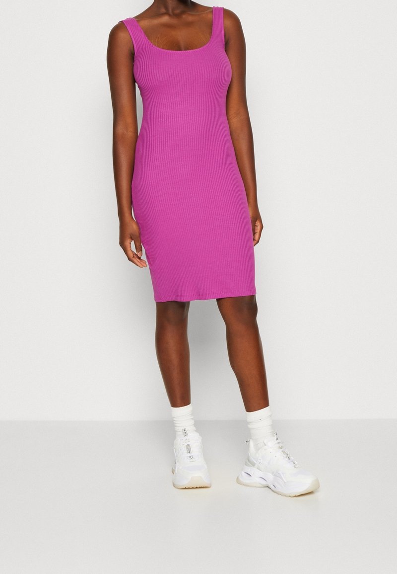 Femme portant une robe violette côtelée sans manches, longueur genou, associée à des chaussettes blanches et des baskets blanches épaisses, debout devant un fond uni.