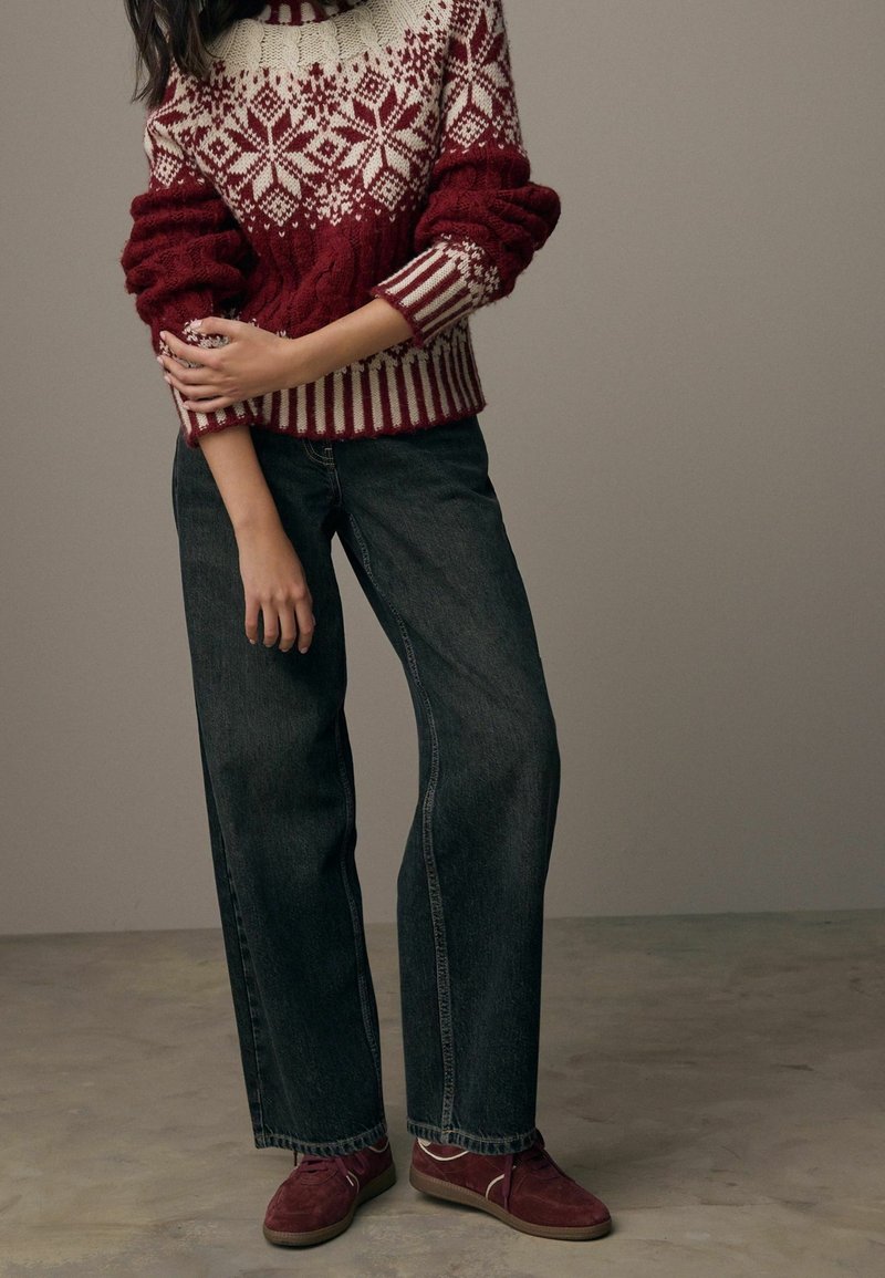 Personne portant un pull rouge et blanc à motifs, un jean large foncé et des baskets rouges, debout les bras croisés sur le torse.