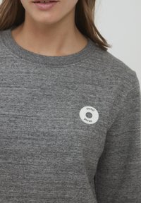 Grå hettegenser med teksturert overflate, med et sirkulært hvitt logo-merke på venstre brystområde. Rund hals-design.