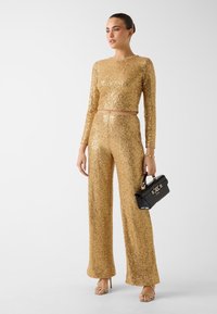 Goldglänzendes zweiteiliges Outfit mit langen Ärmeln und weitem Hosenbein. Kombiniert mit einer schwarzen Handtasche mit goldener Hardware.