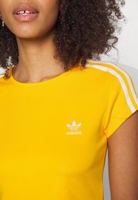 Žluté tričko Adidas s krátkým rukávem, zdobené bílými třemi pruhy na ramenech a malým bílým logem na hrudi.