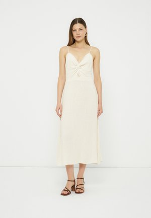 Kvinde iført en ærmeløs, cremefarvet midi-kjole med en rynket frontudskæring og brune, remme hælede sandaler stående mod en hvid baggrund.