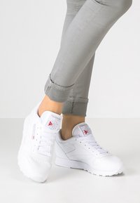 Fötter i vita Reebok Classic sneakers och ljusgrå uppvikta byxor mot en enfärgad ljus bakgrund.