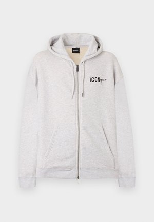 Young man wearing light gray zip-up hoodie, distressed blue jeans, and red sneakers, standing with one hand in pocket, looking to side.