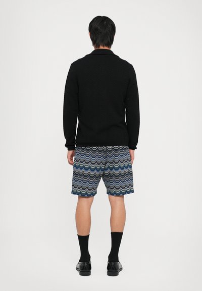 Melnā teksturētā džemperis ar garām piedurknēm, apvienots ar rakstainām zilām šortiem ar viļņotām svītrām; izskatu noslēdz melnas zeķes un apavi.