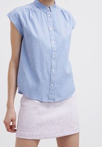Frau in einem hellblauen kurzärmeligen Hemd mit Knopfleiste und einem strukturierten hellrosa Minirock vor einem schlichten weißen Hintergrund.