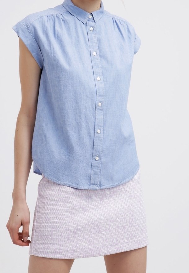 Frau in einem hellblauen kurzärmeligen Hemd mit Knopfleiste und einem strukturierten hellrosa Minirock vor einem schlichten weißen Hintergrund.