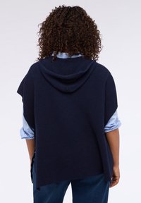 Granatowy ponczo z kapturem, wykonany z fakturowanego materiału, charakteryzuje się luźnym krojem i bocznymi rozcięciami. Noszony na jasnoniebieskiej koszuli i ciemnych dżinsach.